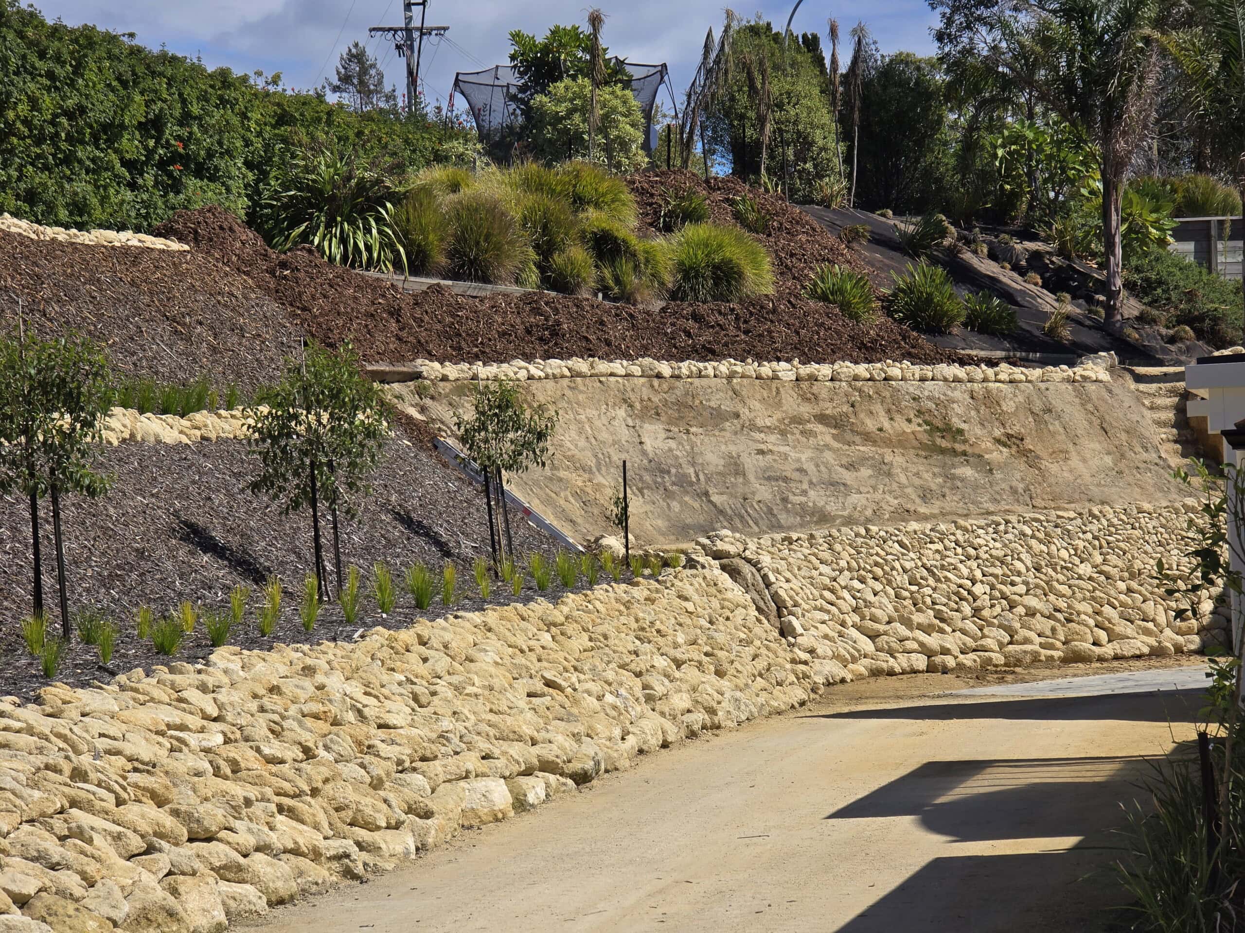 Multiple layers of rock and soil form a landscaped slope with retaining walls, trees, and shrubs in a garden setting.
