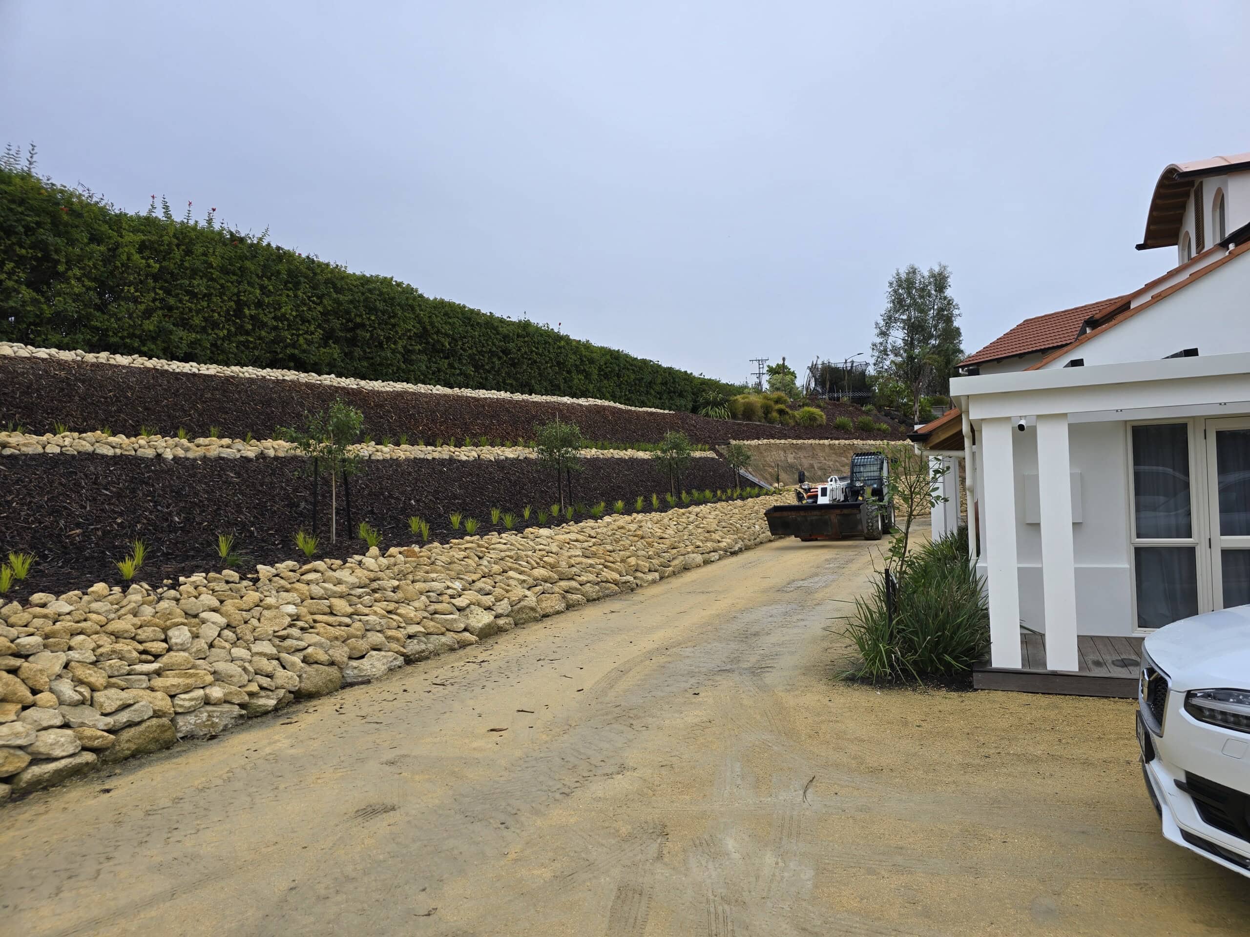 A gravel driveway beside a white house with a stone retaining wall and a bulldozer in the background.