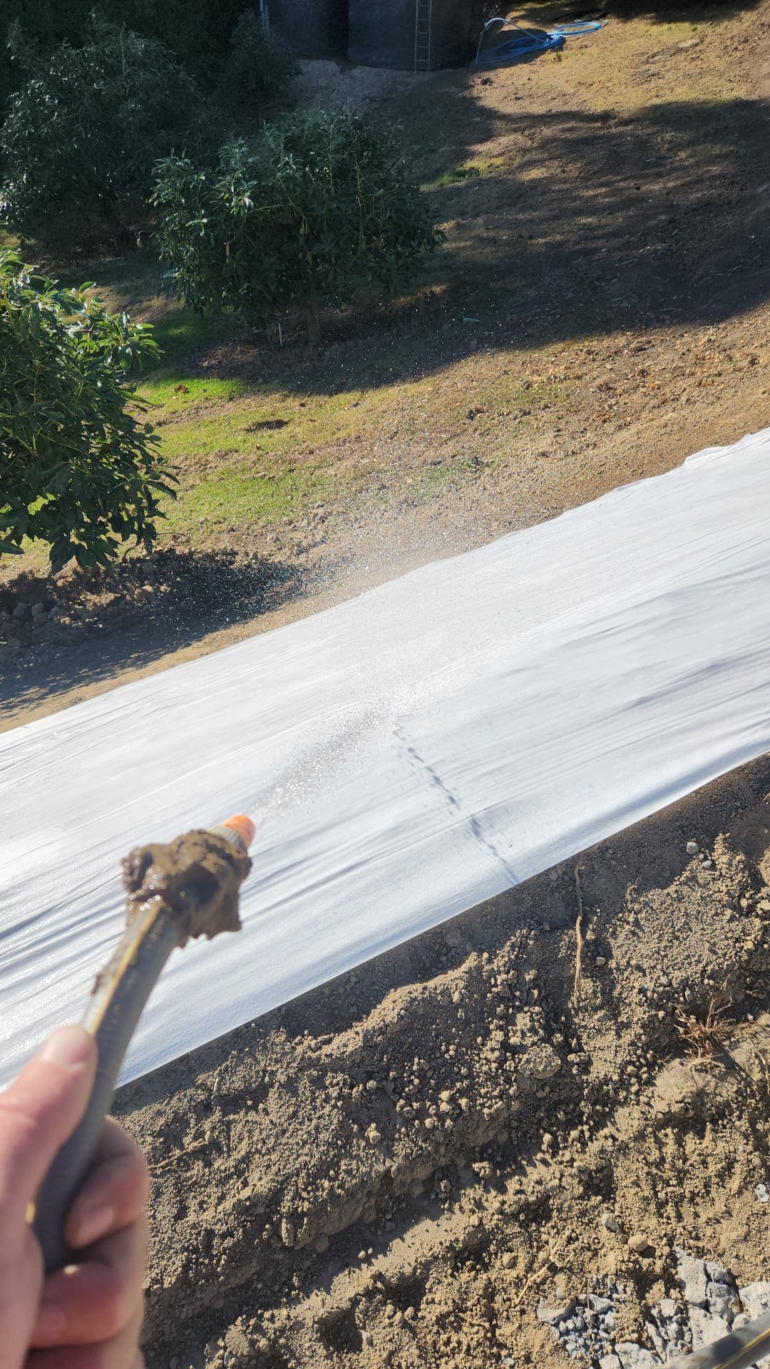 Person spraying a white waterproof membrane on a sloped surface outdoors, with trees and grass in the background.